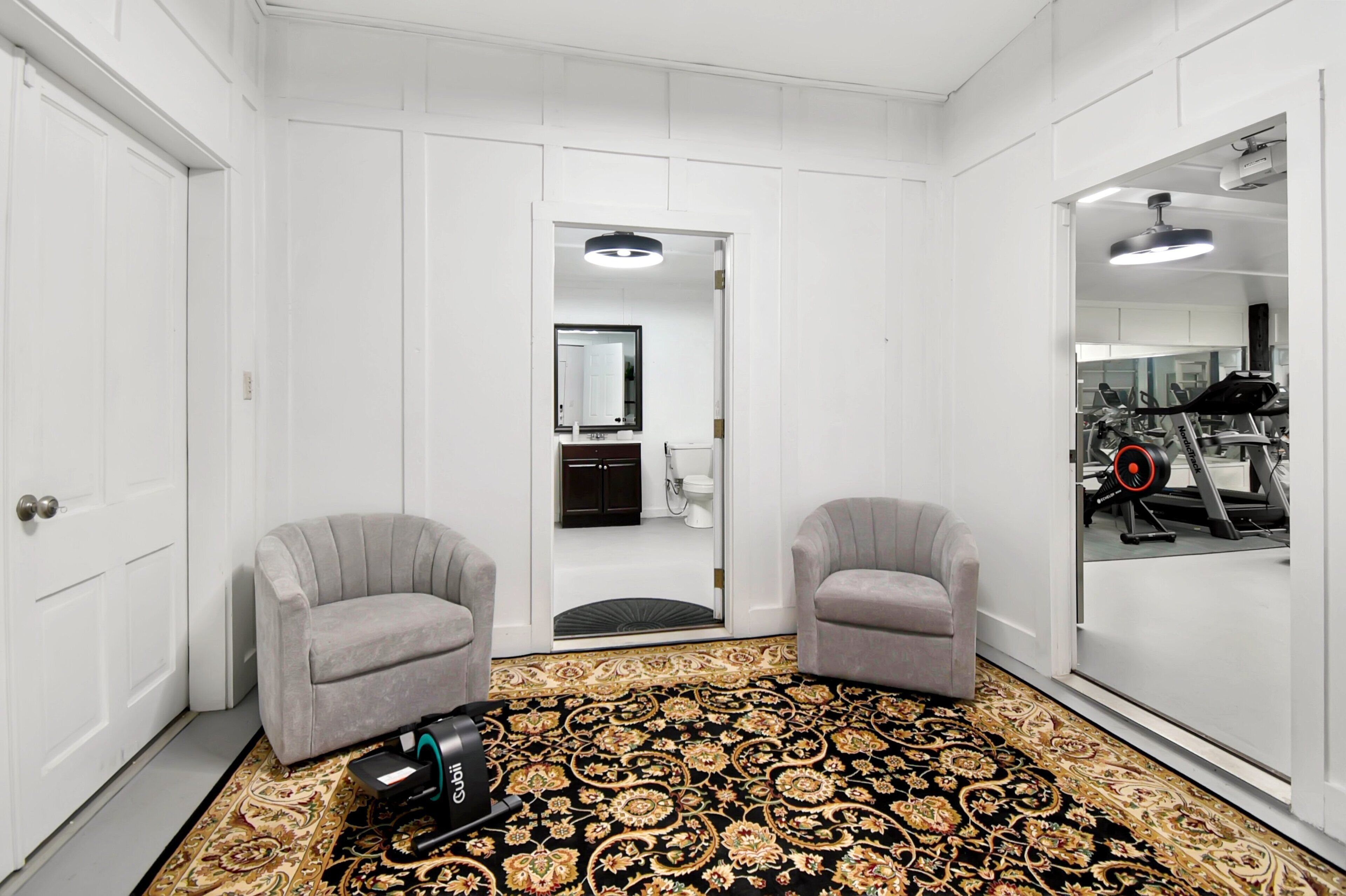 Una habitación con alfombra estampada, dos sillones, una aspiradora, un espejo y vistas a un gimnasio a través de una puerta de vidrio.