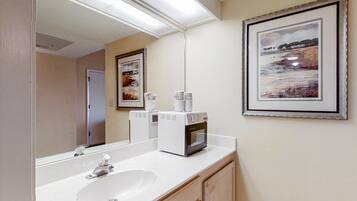 A bathroom with a white sink, a mirror, a framed painting, and a coffee maker.