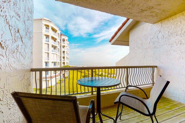 Un balcon avec une table ronde et deux chaises, d’où l’on peut admirer un immeuble et une pelouse verdoyante.
