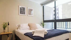 A neatly made bed with a white and dark blue blanket, a small bedside table with a plant, and two framed botanical prints on the wall.