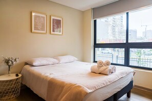 Ein Hotelzimmer mit einem Bett, zwei gerahmten Bildern an der Wand, einer Vase mit Blumen auf einem Beistelltisch und einem Fenster mit Blick auf die Stadt.