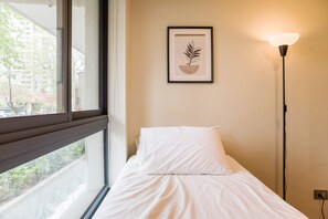 Ein Hotelzimmer mit einem Bett, einem gerahmten Kunstwerk an der Wand, einer Stehlampe und einem Fenster mit Blick auf Grünflächen und Gebäude.