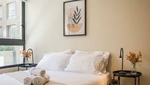Un letto rifatto con lenzuola bianche e asciugamani arrotolati, un comodino con una lampada e un vaso di fiori, e un quadro appeso alla parete.