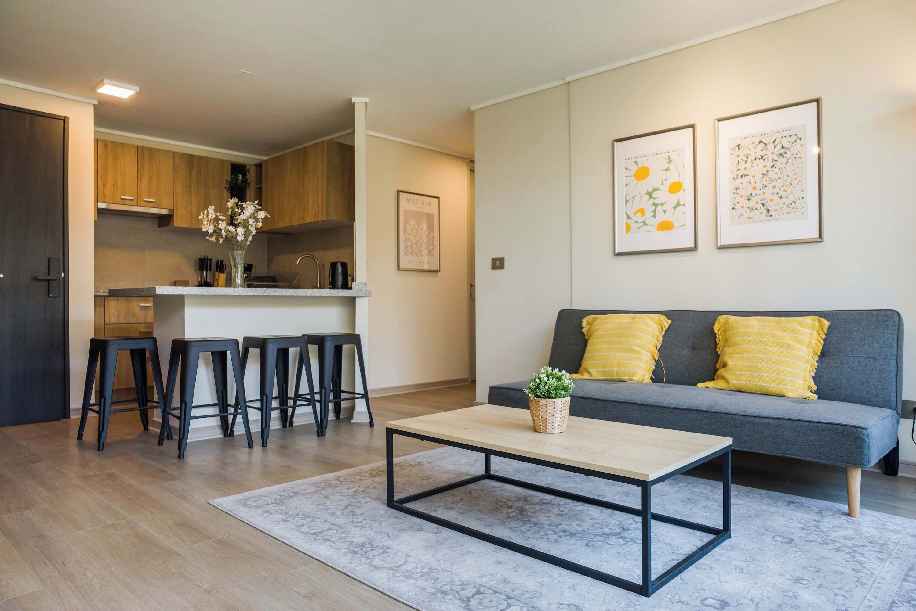 A modern living room with a grey sofa, two yellow cushions, a coffee table, and a kitchen in the background.