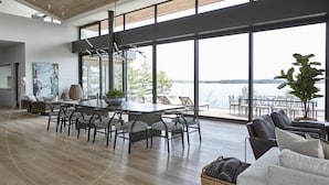 Een ruime woonkamer met een grote eettafel, zachte zithoek, een groot raam met uitzicht op het water, en een moderne keuken op de achtergrond.