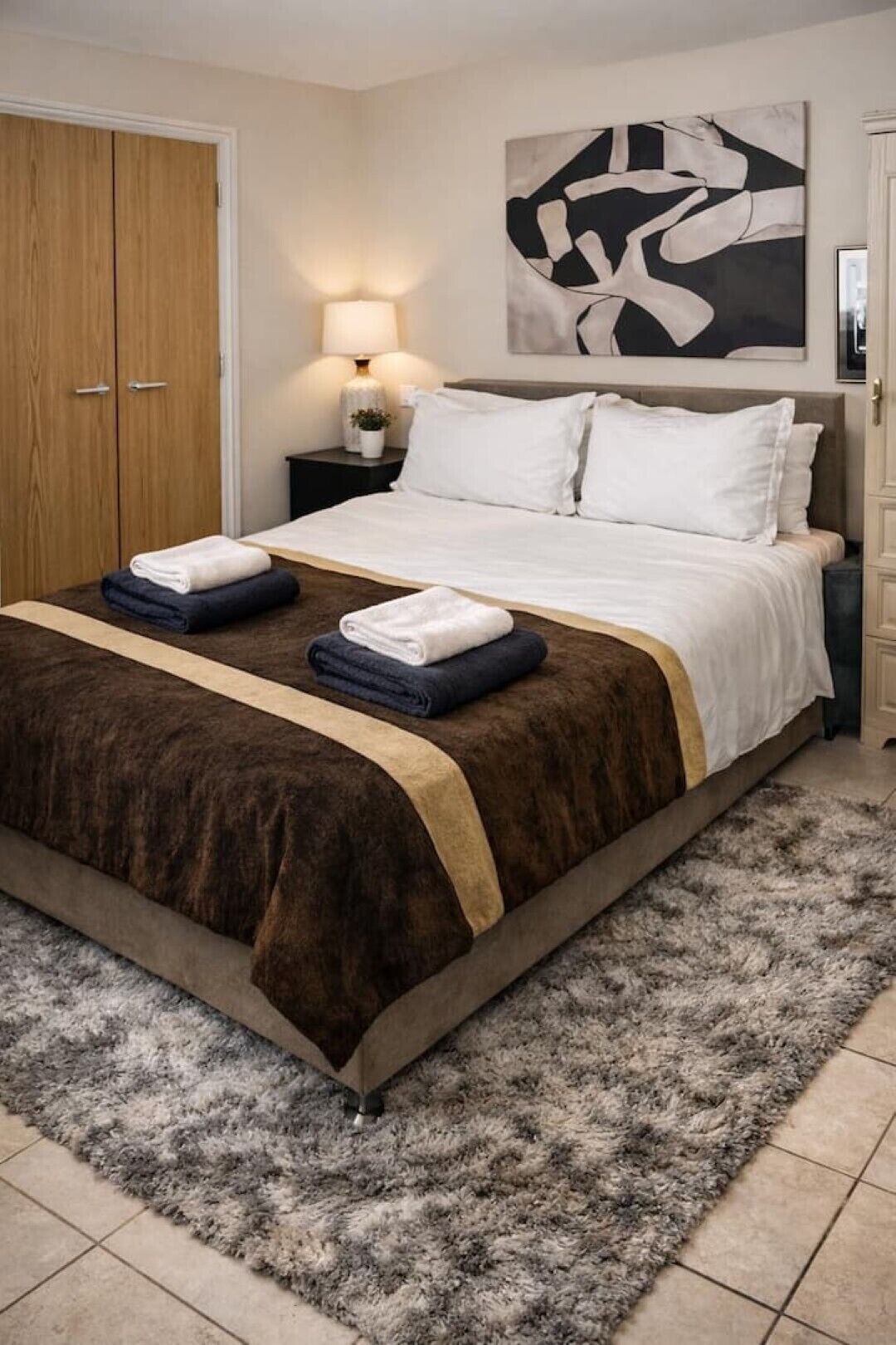 A neatly made bed with a brown and white bedspread, two folded towels, and a fluffy gray rug on a tiled floor.