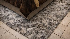 Un lit bien fait, recouvert d’une couvre-lit marron et blanche, avec deux serviettes pliées et un tapis gris moelleux posé sur un sol carrelé.