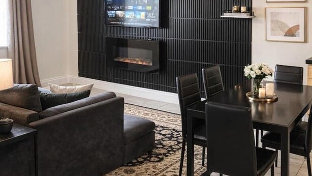Un salon moderne avec un mur d’accent de couleur foncée, un téléviseur à écran plat, une cheminée, un coin repas avec une table et des chaises, et un tapis à motifs.
