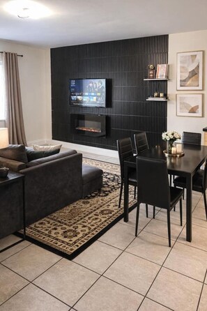 Una sala moderna con una pared de acento oscura, un televisor de pantalla plana, chimenea, zona de comedor con mesa y sillas, y alfombra estampada.