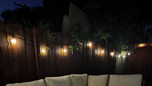 A sectional sofa with a low coffee table, set on a patterned rug, against a wooden fence with string lights. - Boho Vibrant San Diego House w/Patio, Grill & Games (San Diego)