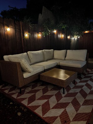 Un sofá modular con una mesita de centro baja, sobre una alfombra estampada, frente a una cerca de madera con luces colgantes.