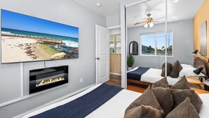 A modern bedroom with a large TV displaying a beach scene, a fireplace, a bed with pillows, and a ceiling fan. - Boho Vibrant San Diego House w/Patio, Grill & Games (San Diego)