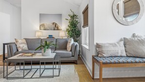 Een moderne woonkamer met een grijze bank, een salontafel en een bijzettafeltje. Er staat een potplant, een spiegel hangt aan de muur, en er is een ingelijste foto.