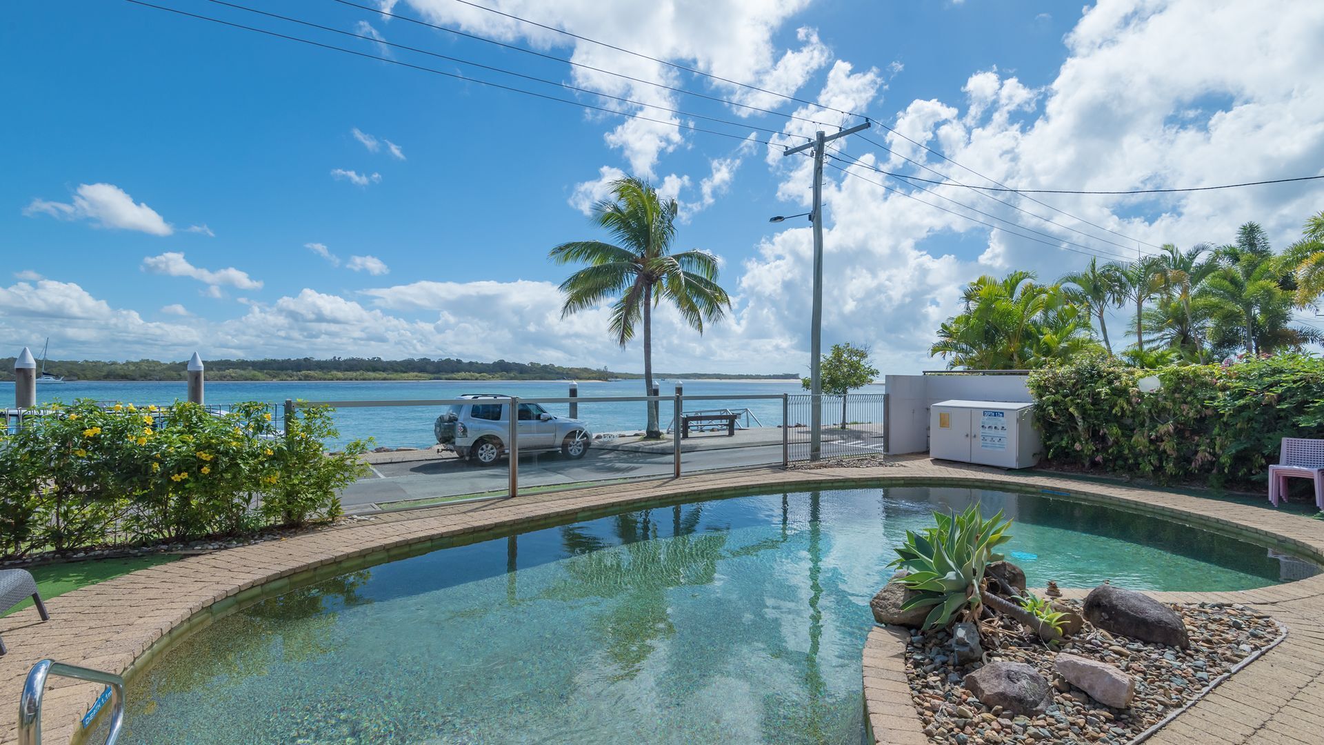 A clear pool with a view of the sea, a parked car, and a palm tree.