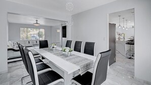 A modern dining room with a long white table, black chairs, and a chandelier.