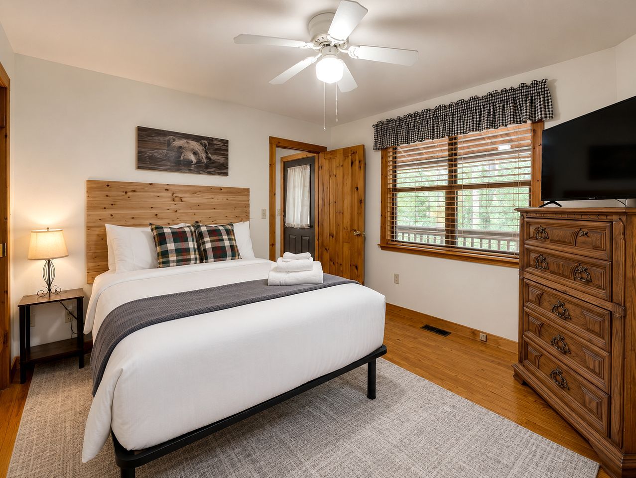 A bedroom with a bed, a wooden dresser, a ceiling fan, a window with blinds, and a wall-mounted TV.