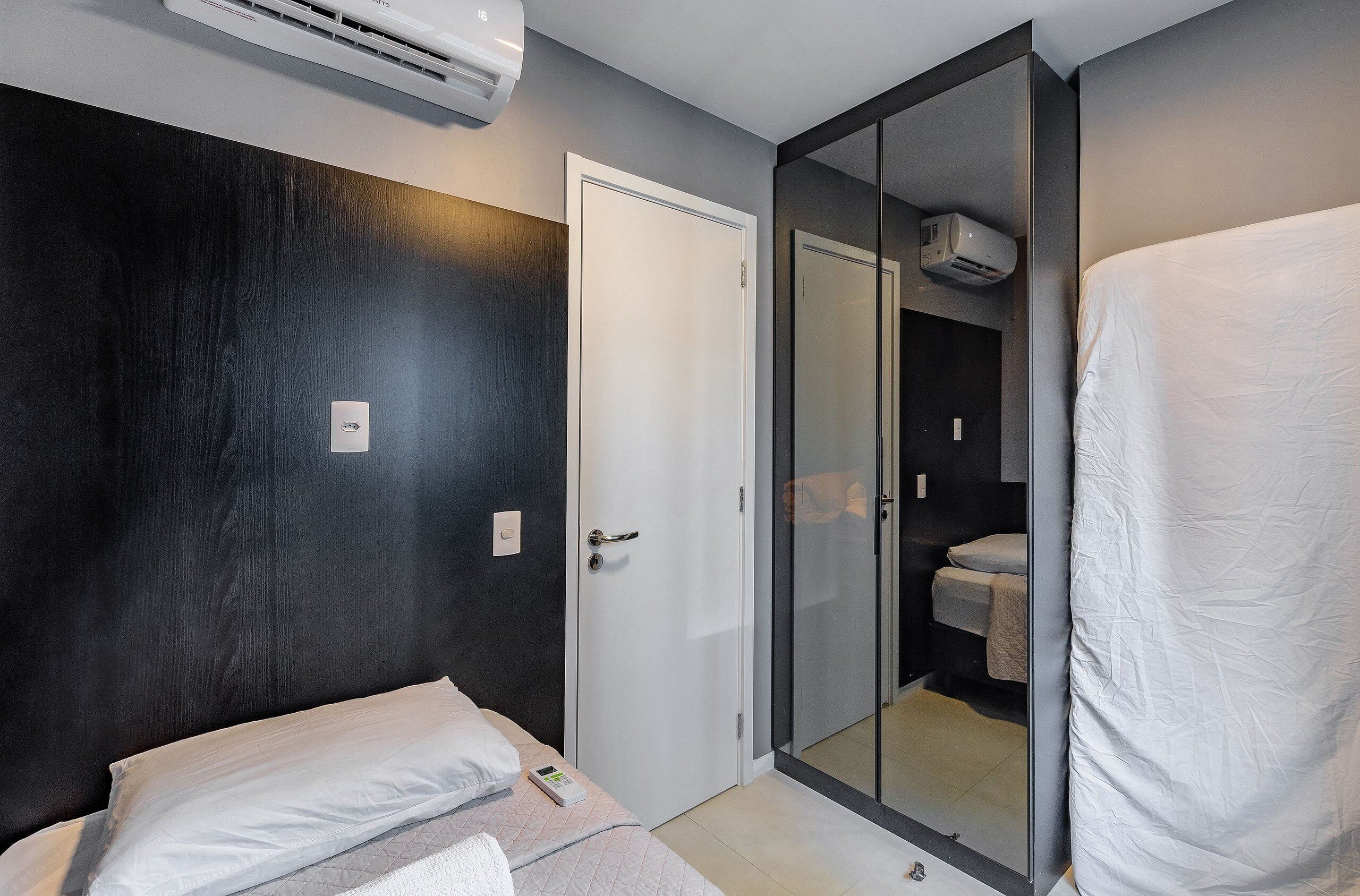 Un dormitorio con cama, armario y baño visible a través de un vidrio. También se observa un equipo de aire acondicionado en la pared.