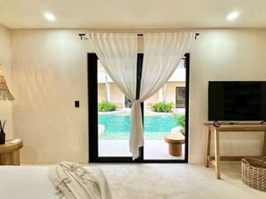 A bedroom with a bed, a TV, a small table, and a view of an outdoor swimming pool. - COCAY PI 2201 Swim Up, Escapada Romantica (Tulum)