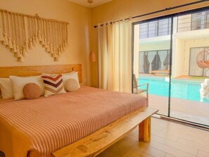 Uma cama grande com cabeceira de madeira, vista de uma piscina através de uma porta de vidro e uma parede com uma franja decorativa.