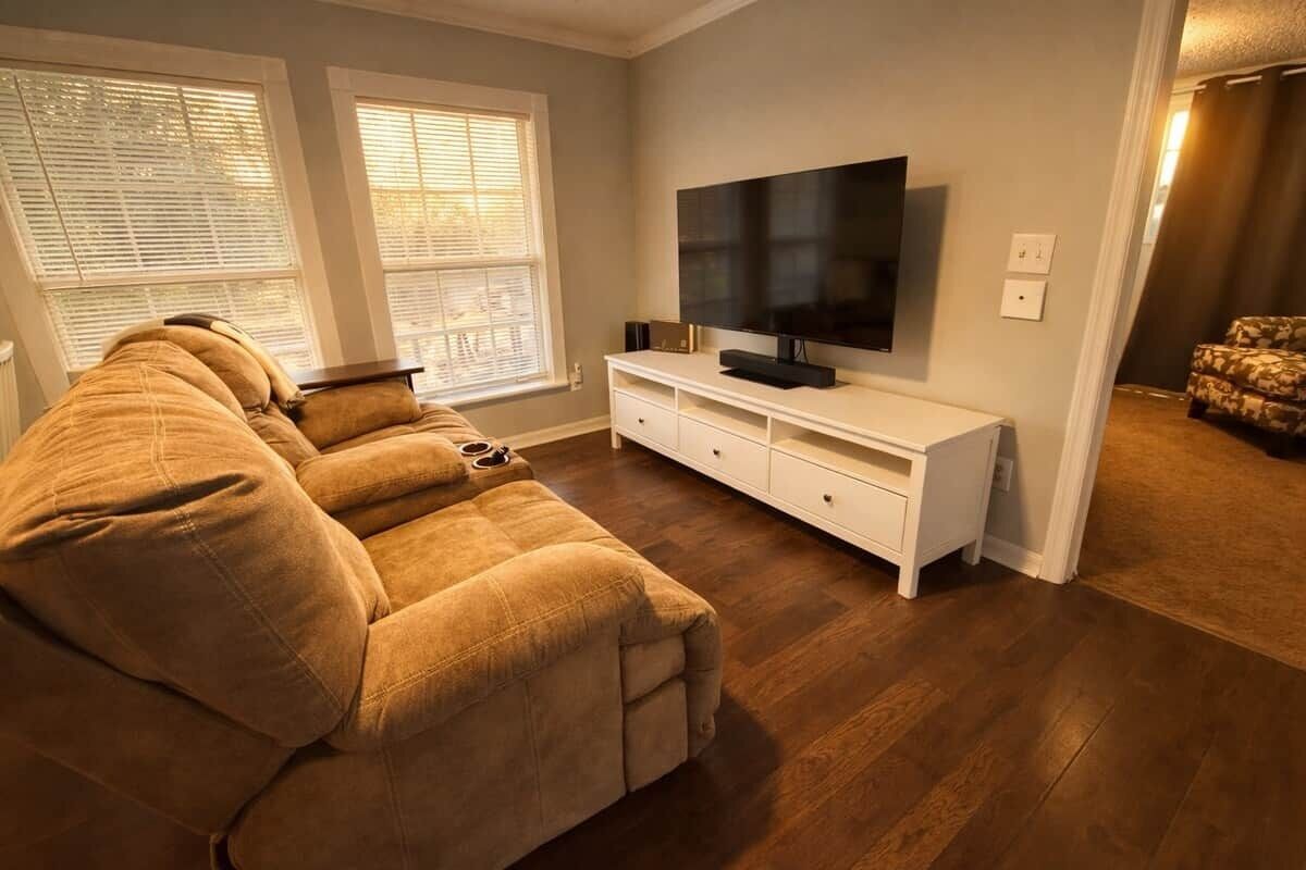Uma sala de estar com um sofá grande, uma televisão de tela plana em um suporte branco e uma janela com persianas.