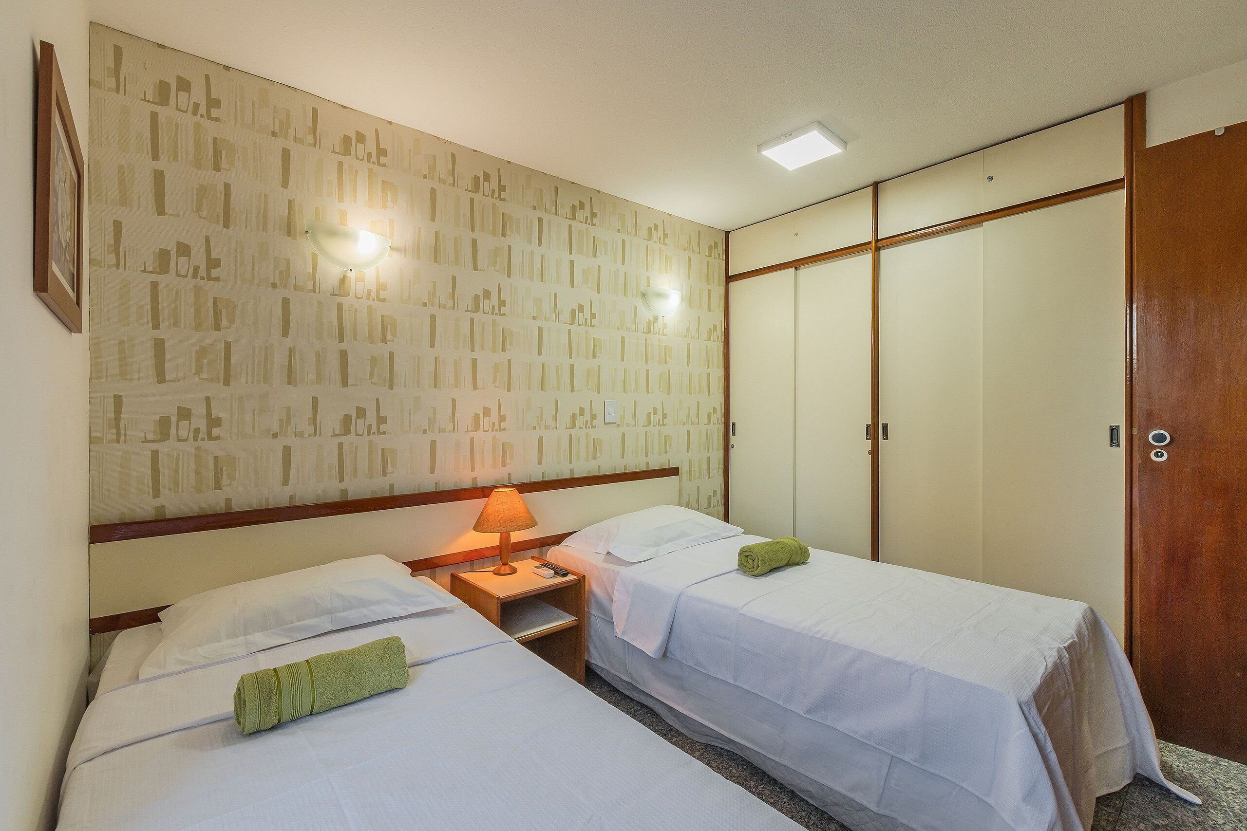 A hotel room with two beds, a nightstand with a lamp, a wardrobe, and a patterned wallpaper.