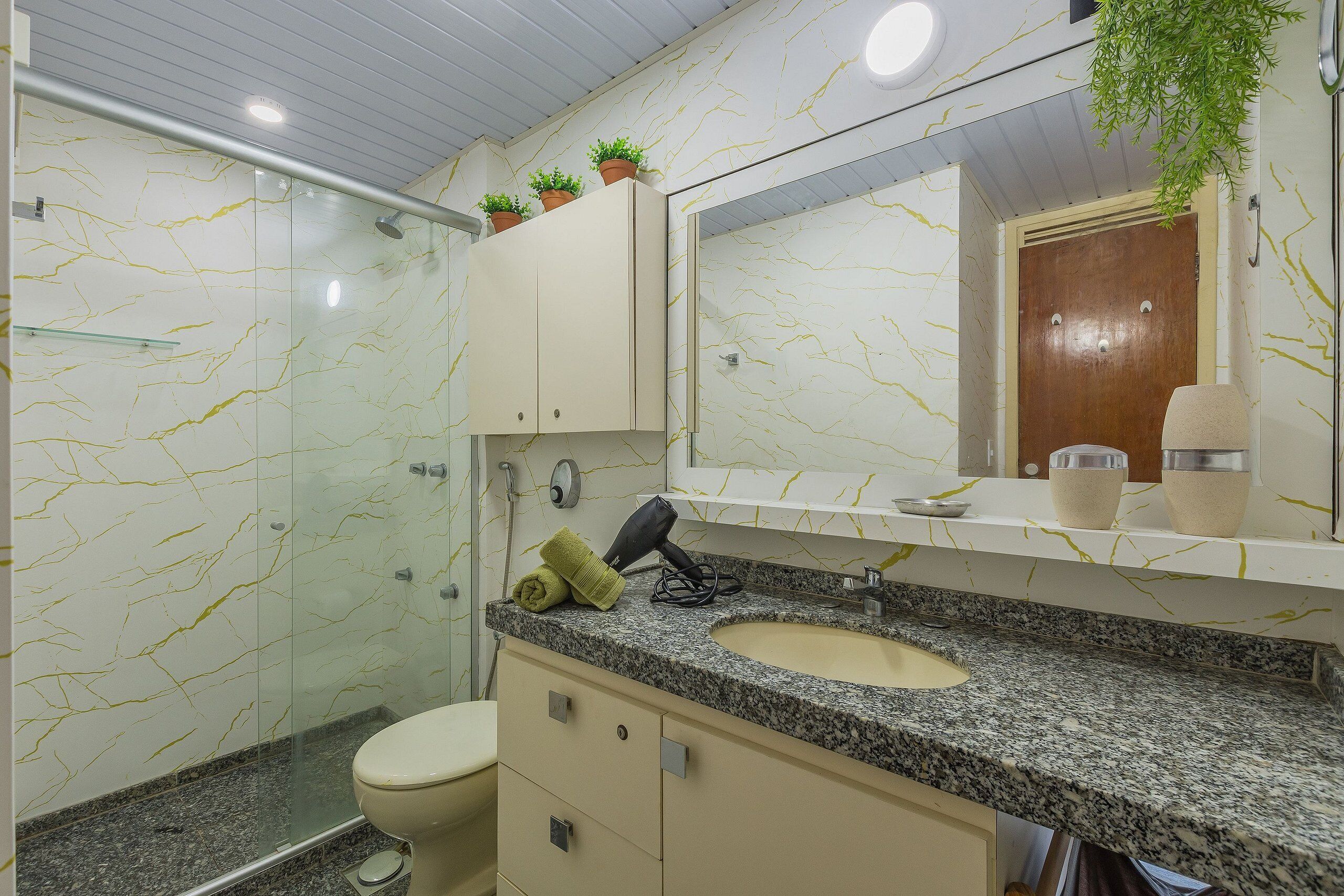 A bathroom with a marble-patterned wall, a glass shower enclosure, a toilet, a sink with a granite countertop, and a mirror above the sink.
