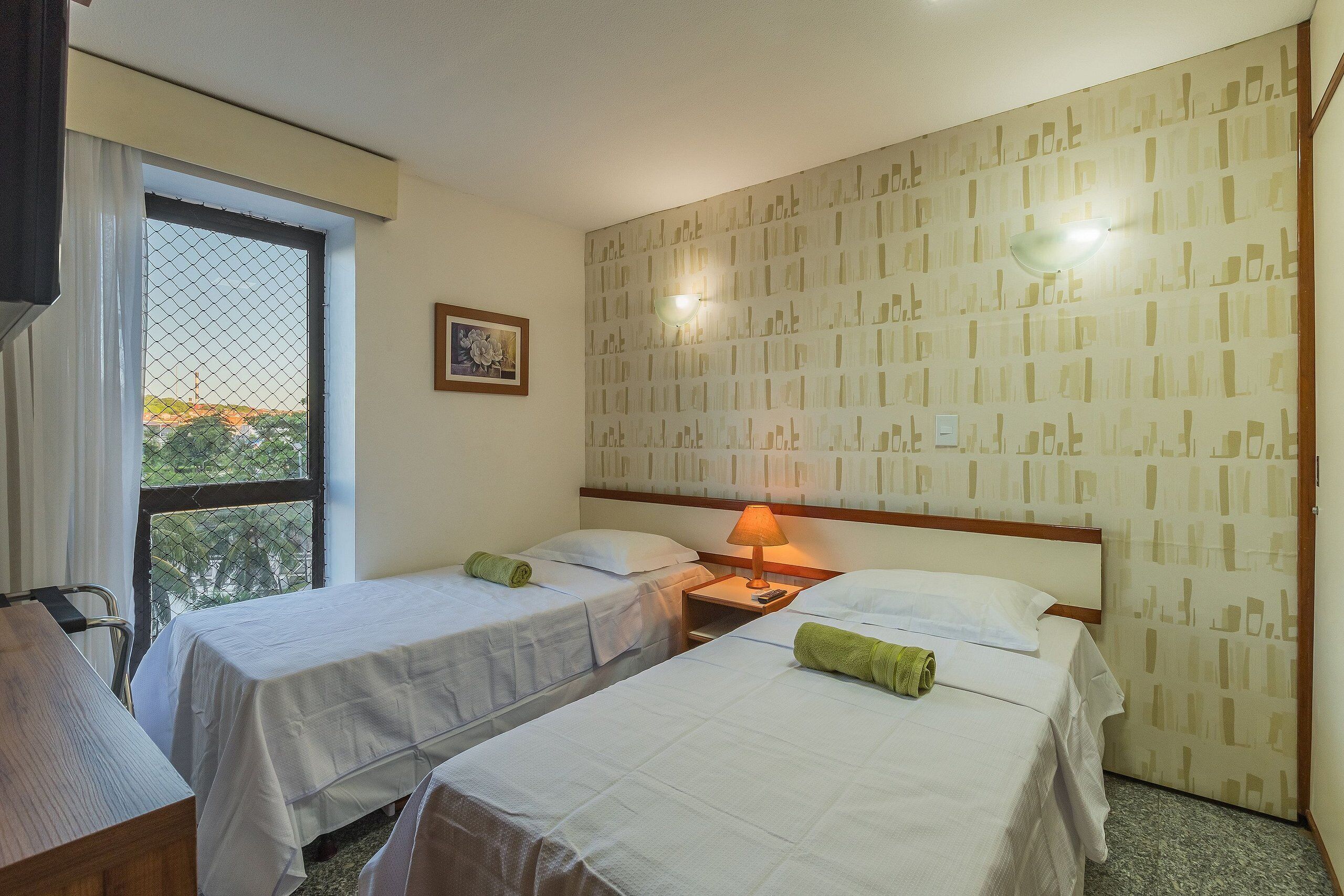 A hotel room with two single beds, a desk, a chair, a window with a view, and a patterned wall.