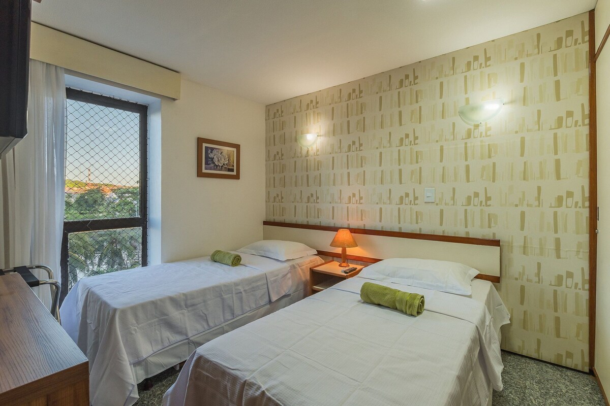 Quarto de hotel com duas camas de solteiro, uma escrivaninha, uma cadeira, uma janela com vista e parede com padrão.