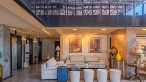 A modern lobby with a high ceiling, a seating area with a sofa and ottomans, a blue suitcase, and artwork on the walls. - Iracema Residence #103 - Seafront by Carpediem (Fortaleza)