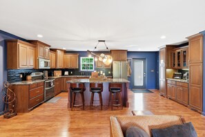 A modern kitchen with wooden cabinets, a central island with bar stools, stainless steel appliances, and a hanging light fixture. - The Grand Ole Hideaway- Mountain Views/Private/Fire Pit/Game Room/Porch/Peaceful (Gatlinburg)