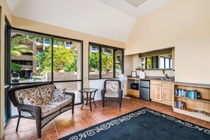 Ein Zimmer mit einer Couch, zwei Stühlen, einem kleinen Tisch, einer Küchenzeile mit Spüle und Mikrowelle sowie großen Fenstern mit Blick ins Grüne.