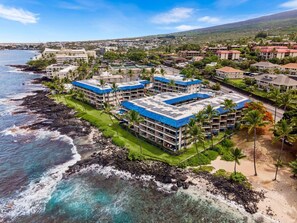 A coastal hotel with multiple buildings featuring blue roofs, situated on a rocky shoreline with clear blue waters. - Kona Reef F-23 (Kailua-Kona)