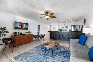 Ein modernes Wohnzimmer mit Sofa, Couchtisch und Essbereich. Es gibt eine Küche mit Bar und Deckenventilator.