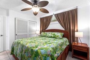 A bedroom with a bed, a ceiling fan, a nightstand, and a window with curtains. - Kona Reef F-23 (Kailua-Kona)