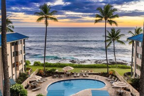 A resort pool with lounge chairs and umbrellas, overlooking the ocean with palm trees and a view of the sea. - Kona Reef F-23 (Kailua-Kona)