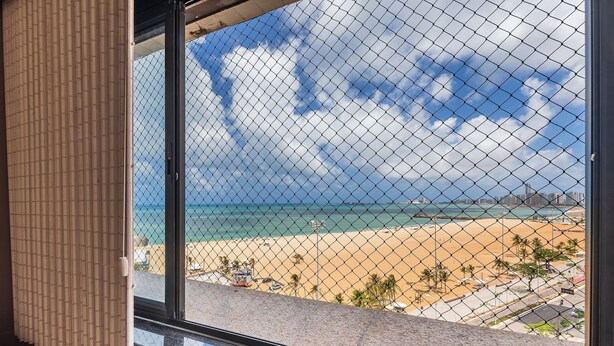 A view through a window with a chain-link fence, featuring a beach, ocean, and distant buildings. - Aquidaba #803 - Apto With sea View by Carpediem (Fortaleza)