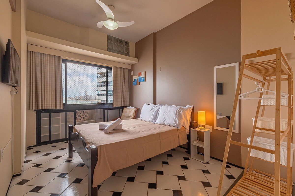 A hotel room with a bed, a ceiling fan, a window with curtains, a wooden shelf, and a mirror.