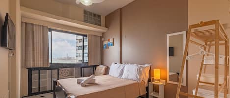 A hotel room with a bed, a ceiling fan, a window with curtains, a wooden shelf, and a mirror.
