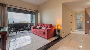 A modern living room with a red sofa, a glass table, and chairs. There is a window with curtains, a lamp, and a view of the city. - Aquidaba #803 - Apto With sea View by Carpediem (Fortaleza)