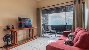 A living room with a TV on a wooden stand, a red sofa, a fan, and a balcony with chairs. - Aquidaba #803 - Apto With sea View by Carpediem (Fortaleza)