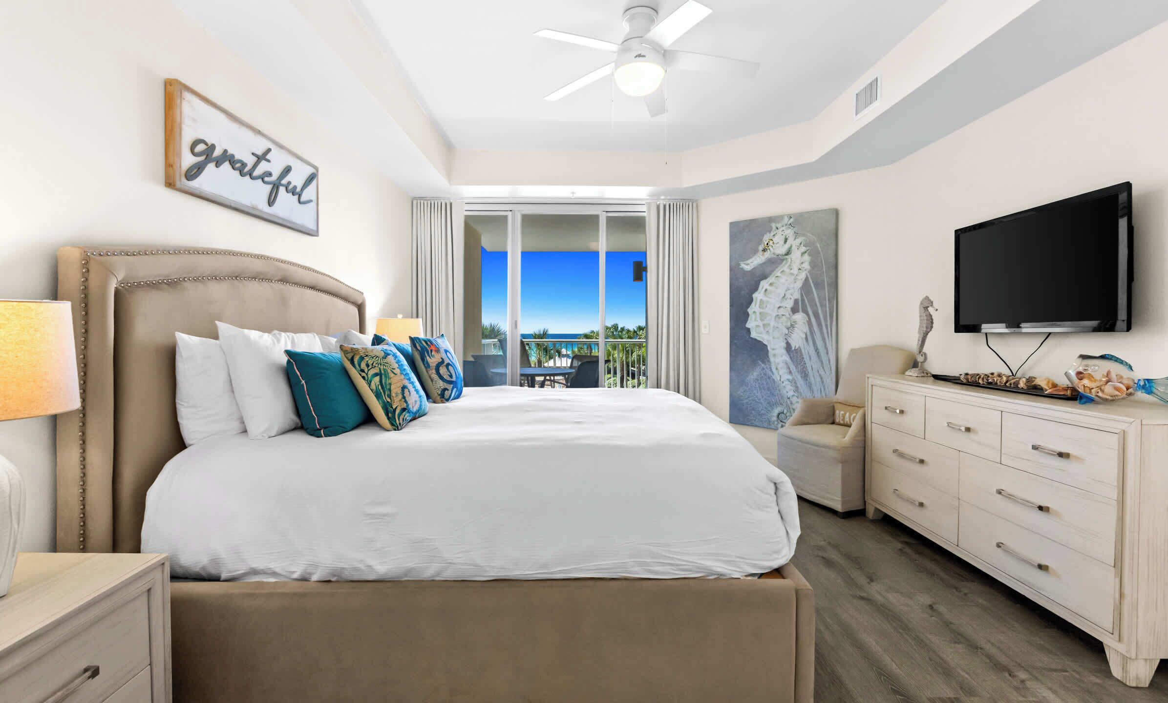 A bedroom with a bed, a TV, a ceiling fan, a painting, and a view of the ocean.
