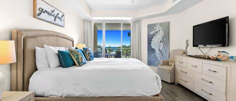 A bedroom with a bed, a TV, a ceiling fan, a painting, and a view of the ocean.