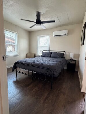 A bedroom with a bed, a ceiling fan, two windows with blinds, an air conditioning unit, and a nightstand with a lamp. - Hooked On Bay Views Palacios (Palacios)