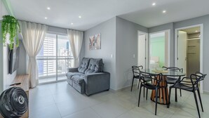 Ein modernes Wohnzimmer mit grauem Sofa, einem Esstisch mit Stühlen, einem Ventilator und einem großen Fenster mit Vorhängen.