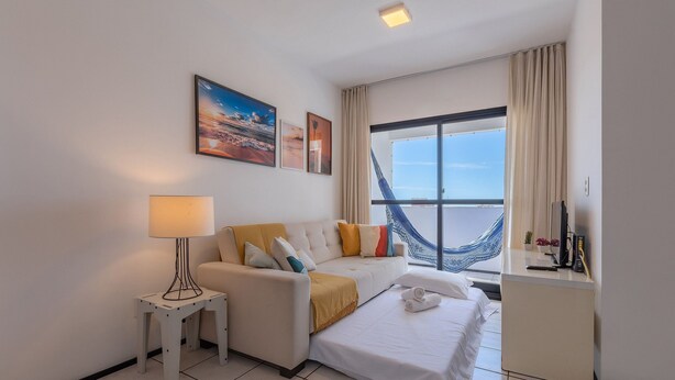 Quarto de hotel com cama, sofá, mesinha com abajur, escrivaninha com computador e uma grande janela com vista para o mar.