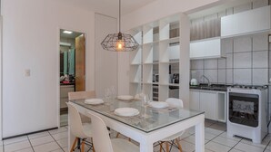 Uma cozinha moderna com armários brancos, forno embutido e área de jantar com mesa de vidro e cadeiras brancas.