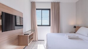 Quarto de hotel moderno com televisão de tela plana na parede, um aparador de madeira, cama bem arrumada com lençóis brancos, janela com cortinas bege e mesa de cabeceira com luminária.