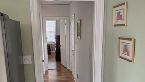 Un pasillo con suelo de madera, paredes blancas y una puerta que da a otra habitación. Hay cuadros en la pared y un refrigerador a la izquierda.