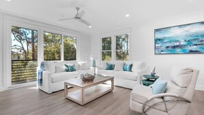 Een ruime woonkamer met een witte zithoek, een salontafel en een fauteuil. Grote ramen zorgen voor natuurlijk licht en uitzicht op bomen.