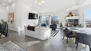 Een moderne woonkamer met een witte bank, een ronde eettafel met stoelen, een aan de muur bevestigde televisie en een kerstboom.